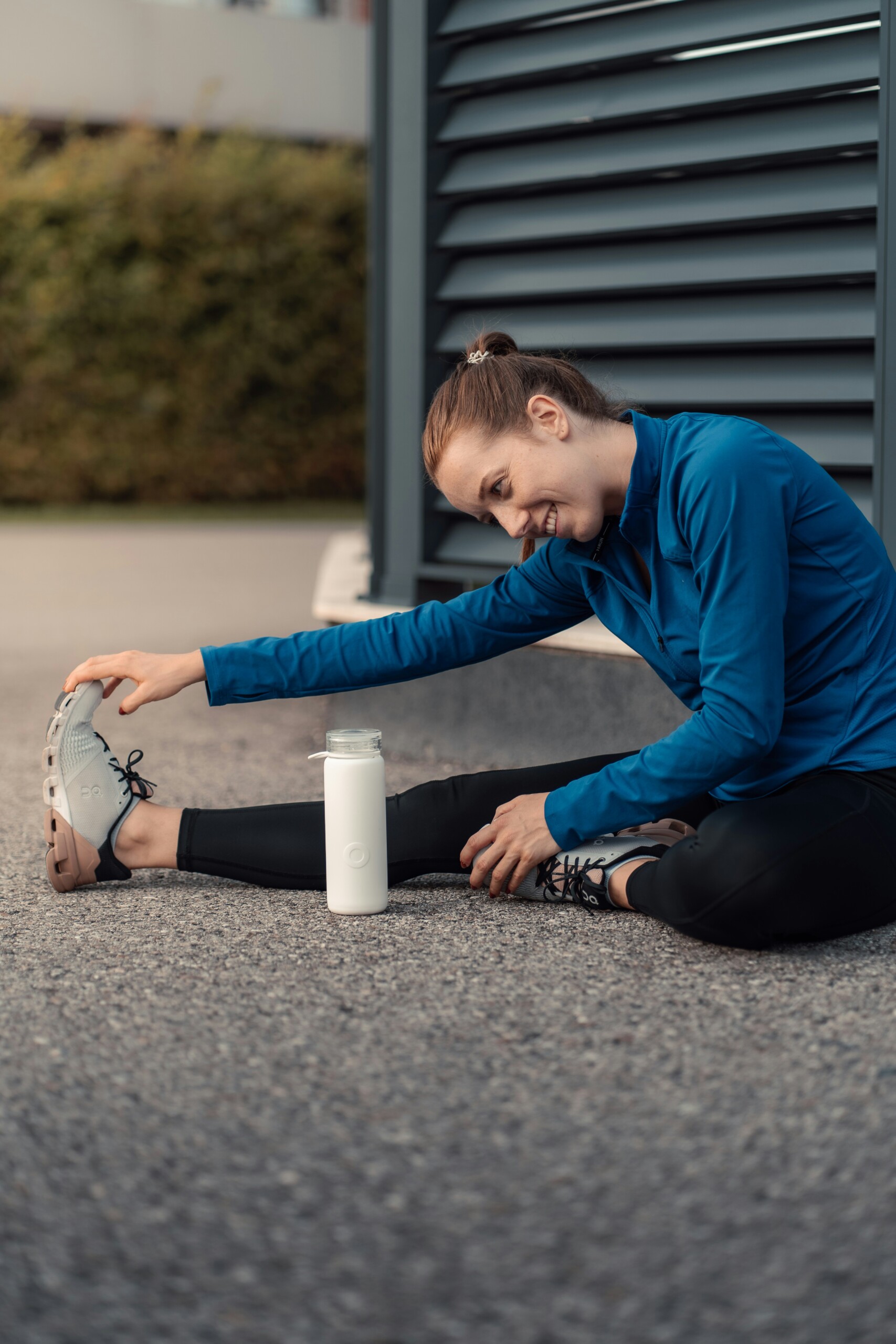 girl in black leggings and blue fitness jacket on the ground stretching with waterbottle beside her